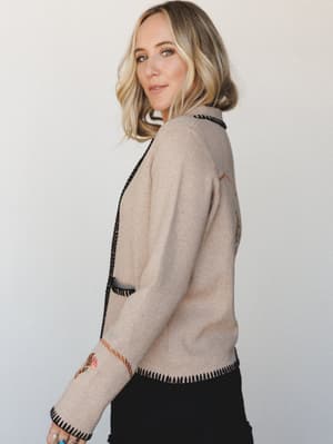 A blonde model shot in profile against a clean white wall, wearing a oatmeal knit cardigan with black whipstitch trim and subtle embroidery — the kind of piece that looks vintage but feels brand new.