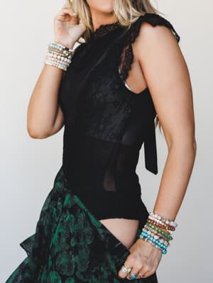 A cropped side-angle shot of a black lace and mesh bodysuit styled with a dark floral skirt, the focus pulled toward layered beaded bracelets and silver rings stacked on both wrists — moody, romantic, and very much the outfit you wear when you mean i