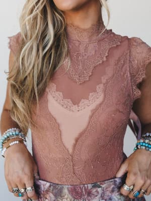 A close-up torso shot of a dusty rose lace bodysuit with intricate scalloped trim and a mock neck, styled with stacked turquoise and beaded bracelets and silver rings over a floral skirt — romantic and a little heirloom.