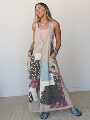 A smiling model wears a wide-leg patchwork jumpsuit in light taupe layered over a blush pink tank, styled with stacked bead bracelets, a delicate silver necklace, and turquoise slide sandals — the whole look reads like a vintage quilt got a really go