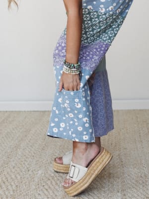 A close-up detail shot of a patchwork midi skirt or wide-leg pants in mixed floral and paisley prints — daisy, teal ditsy, and lavender bandana panels — styled with stacked beaded bracelets and cream platform sandals on a neutral jute rug.