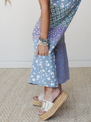 A close-up detail shot of a patchwork midi skirt or wide-leg pants in mixed floral and paisley prints — daisy, teal ditsy, and lavender bandana panels — styled with stacked beaded bracelets and cream platform sandals on a neutral jute rug.