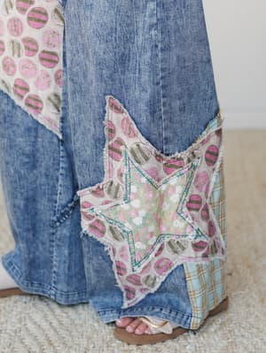 A close-up of wide-leg denim jeans with playful mixed-fabric patchwork at the hem — star appliqué, polka dot panels, floral print, and plaid piecing all stitched together with raw frayed edges for a free-spirited, one-of-a-kind feel.