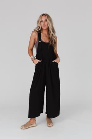 A woman with long wavy blonde hair wears The Nest Serenity Wide Leg Jumpsuit - Black, colorful bracelets, and beige sandals, standing against a white background with her hands in her pockets.
