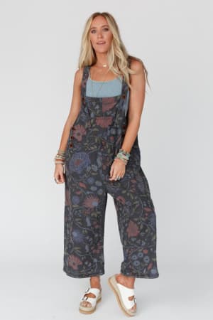 A woman with long blonde hair wears the High Hopes Slouchy Denim Overalls in Floral Black Denim, paired with a blue tank top, stacked bracelets, and white platform sandals, standing against a plain light gray background.