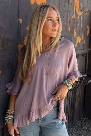 A woman with long blonde hair wears The Nest Let Me Be Ruffled Top in Pale Plum, paired with denim jeans and layered jewelry, as she stands against a weathered dark wooden wall, gazing to the side.