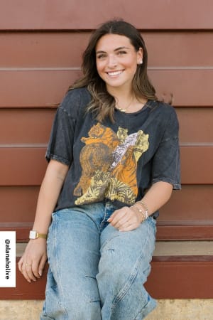 A woman with long brown hair, wearing the On The Prowl Distressed Graphic Tee in charcoal and blue jeans, sits and smiles at the camera in front of a brown wooden wall.