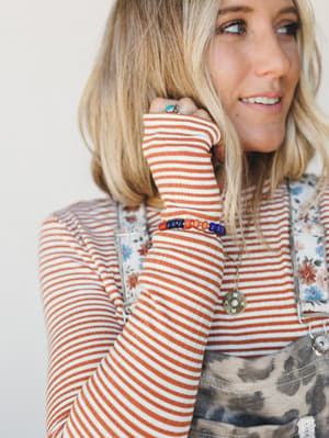 A close-up lifestyle shot of a model layering a rust-striped ribbed long sleeve under a floral and leopard print overall dress, with the beaded bracelet — orange and cobalt blue — front and center on her wrist alongside a turquoise ring and a medalli