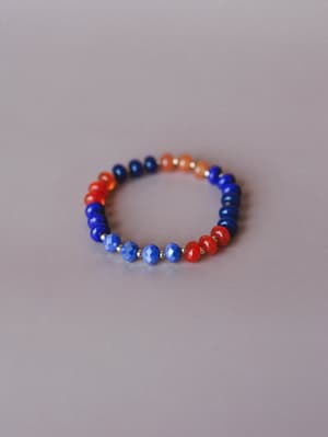 A beaded stretch bracelet laid flat on a muted gray surface — cobalt blue faceted stones mix with red-orange and peach beads, punctuated by small gold spacers for a bold, color-blocked feel.