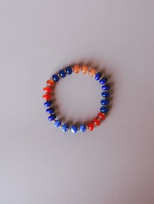 A color-blocked beaded stretch bracelet laid flat — cobalt blue and red-orange faceted stone beads alternate in bold contrast, separated by small gold spacers, shot clean against a soft grey background.