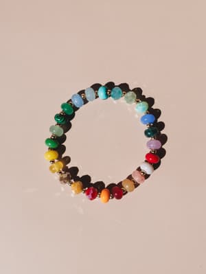 A rainbow-spectrum beaded stretch bracelet laid flat on a warm blush surface, catching natural light — every bead a different color and texture, like someone collected tiny gems from everywhere they'd ever been.