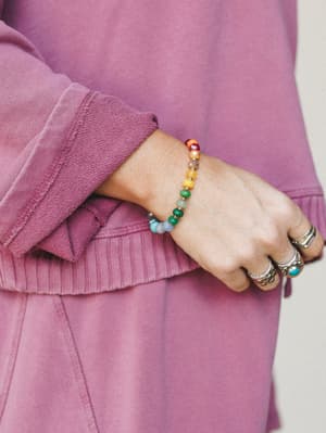 A close-up of a wrist stacked with silver rings and a multicolor beaded bracelet — all resting against the ribbed cuff of a washed mauve sweatshirt, giving the whole thing that effortlessly collected, everyday-magic kind of feel.