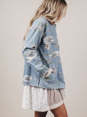 Woman wearing a floral denim jacket over a white dress with a beige background