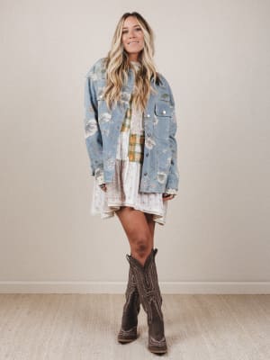 Woman wearing a floral denim jacket, plaid shirt, and white skirt with brown cowboy boots on a plain background