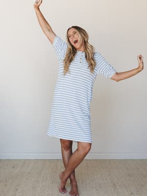 A laughing, arm-raised model dances barefoot in a relaxed striped t-shirt dress with a henley neckline — the kind of dress that makes you feel like it's already Saturday morning.