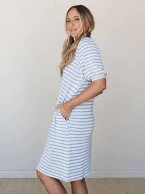 A relaxed striped knit midi dress in soft sky blue and cream, worn with hands tucked into side pockets — the kind of dress that feels like a cozy day off but looks intentional enough for a farmers market run.