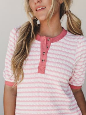 A close-up of a pink-striped henley top in a cozy textured knit — the kind of piece that reads effortlessly sweet, styled with playful pigtails and shot against a soft neutral backdrop.