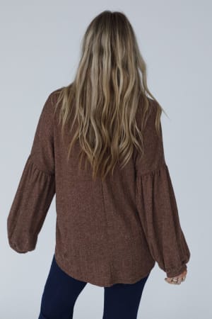 A woman with long, wavy blonde hair stands with her back to the camera, wearing The Nest Sweet Heart Balloon Sleeve Knit Sweater in mocha and dark pants against a plain, light-colored background.