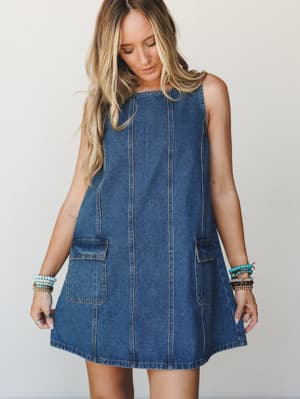 A sleeveless denim mini dress with a relaxed A-line silhouette and front patch pockets, styled with stacked beaded bracelets for an easy, free-spirited everyday look.