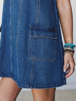 A close-up detail shot of a medium-wash denim mini dress with a large cargo-style patch pocket, styled with a stack of beaded bracelets that bring a relaxed boho energy to the otherwise casual silhouette.