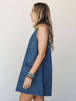 A relaxed denim mini dress with an A-line swing silhouette, shot in profile against a clean studio wall — styled with stacked turquoise and beaded bracelets that give it that effortless boho-everyday energy.