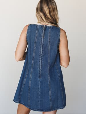 A back view of a sleeveless denim mini dress with a relaxed A-line silhouette, shown on a model against a neutral studio backdrop — the kind of effortless piece that works just as hard on a weekend errand run as it does tucked into a summer memory.