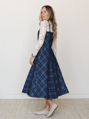 A navy plaid midi overall dress layered over a long-sleeve lace top — the kind of outfit that feels equally at home at a fall market or a casual dinner, styled with woven flats and a stack of turquoise bracelets.