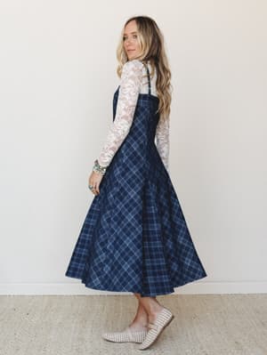 A navy plaid midi overall dress layered over a long-sleeve lace top — the kind of outfit that feels equally at home at a fall market or a casual dinner, styled with woven flats and a stack of turquoise bracelets.