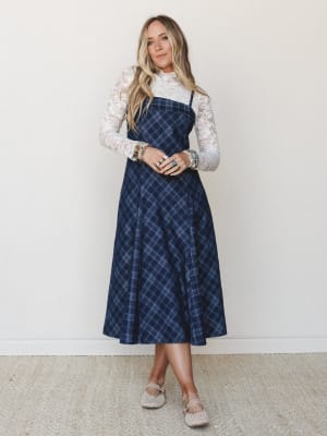 A navy plaid midi slip dress layered over a long-sleeve floral lace top, styled with woven flats and a stack of mixed bracelets — the kind of outfit that feels equally at home at a fall farmers market or a casual holiday dinner.