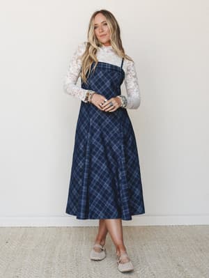 A navy plaid midi slip dress layered over a long-sleeve floral lace top, styled with woven flats and a stack of mixed bracelets — the kind of outfit that feels equally at home at a fall farmers market or a casual holiday dinner.