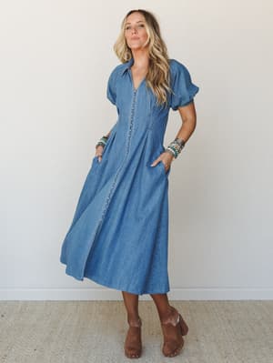 A full-length denim midi dress with puff sleeves and a fit-and-flare silhouette, styled with stacked boho bracelets and brown suede clogs against a clean studio backdrop — the kind of dress that makes a farmer's market run feel like a whole moment.