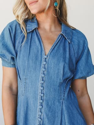 A close-up of a soft denim button-front dress with covered buttons and puff sleeves, styled with gold hoop and turquoise drop earrings against a clean studio backdrop — effortless and a little vintage in the best way.