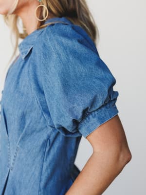 A close-up detail shot of a medium-wash denim dress with a beautifully puffed short sleeve — the kind of piece that feels vintage-inspired but totally wearable, styled with gold hoop earrings and a peek of turquoise necklace.