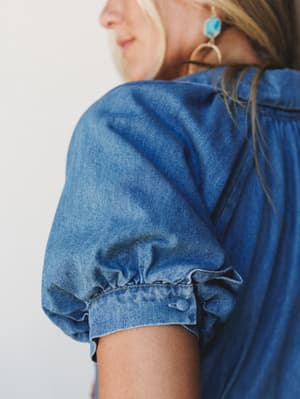 A close-up detail shot highlighting the puff sleeve of a soft chambray denim top or dress, styled with a turquoise drop earring that adds a sweet boho touch.