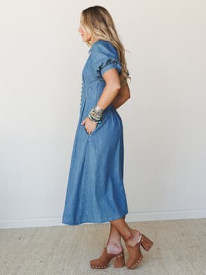 A denim midi-maxi dress with a relaxed A-line silhouette and front button detail, styled with tan suede clogs and a stack of boho beaded bracelets — the kind of outfit that looks effortless but clearly wasn't an accident.