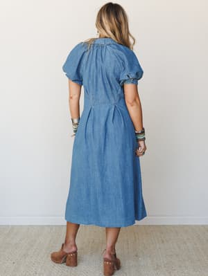 A denim midi dress shot from the back — puff sleeves, a fitted waist with pleating, and an easy A-line skirt that lands below the knee, styled with stacked beaded bracelets and brown platform clogs for a lived-in, effortlessly boho feel.