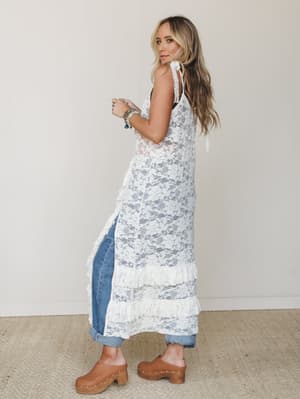 A sheer ivory lace midi duster with tiered ruffle hem is styled over cuffed jeans and wood-heeled clogs — the kind of layered, lived-in look that feels effortlessly put together without trying too hard.