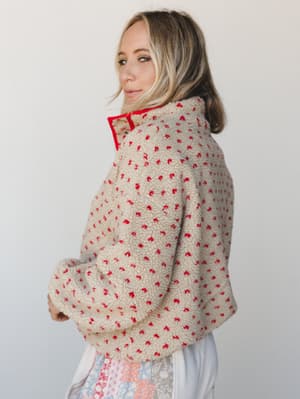 A model shot from the side showing a cozy, oversized sherpa pullover in cream with scattered red floral speckling and a red zip collar — the kind of jacket that makes you want to grab a coffee and go nowhere fast.