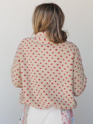 Shot from the back, this cozy sherpa pullover in cream with scattered red floral speckles has that perfectly worn-in, grab-it-off-the-chair energy — layered over a floral skirt and white pants for an effortlessly relaxed look.