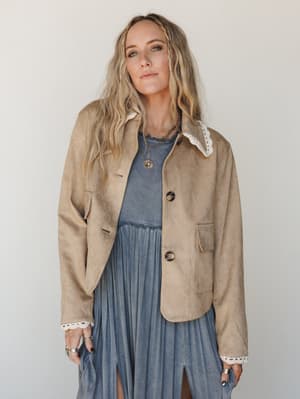 A cropped tan suede-look jacket with cream lace trim at the collar and cuffs, layered over a washed blue pleated midi dress — the kind of outfit that looks like it came together effortlessly but definitely didn't.
