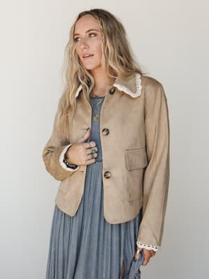 A tan suede-look jacket with cream lace trim at the collar and cuffs layered over a dusty blue maxi dress — the kind of outfit that feels vintage-collected without trying too hard.
