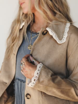 A close-up of a tan suede-look jacket with cream crochet trim at the collar and cuffs, layered over a faded blue top — styled with stacked gold rings and layered necklaces for that perfectly lived-in, effortlessly collected look.