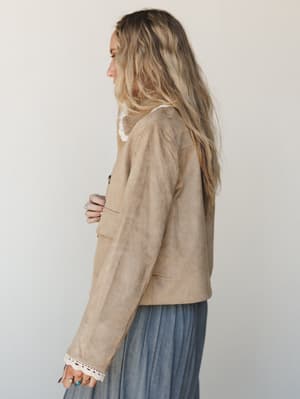 A side-profile shot of a cropped tan suede-look jacket with delicate crochet lace trim at the cuffs and collar, layered over a soft blue pleated skirt — the kind of piece that makes a thrifted-feeling outfit look completely intentional.