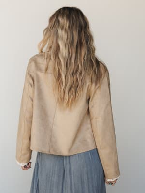 Shot from behind, a model in a cropped tan faux-suede jacket over a dusty blue midi skirt — the back seam detailing and lace-peeking cuffs give it that quiet, personal-style energy that doesn't need to announce itself.