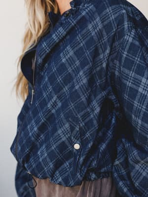 A close-up detail shot of a plaid denim jacket in deep indigo, showing the zip front, snap-button pocket, and ruffled hem — layered over what looks like a soft taupe skirt or dress.