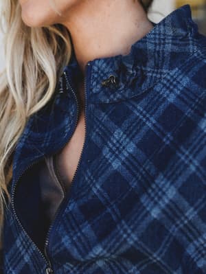 A close-up detail shot of a navy plaid jacket with a zip-front closure and snap-button collar, shot with warm natural light that brings out the texture of the washed denim-like fabric.