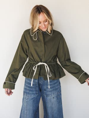 An olive cropped jacket with cream contrast piping and a drawstring waist tie is styled over wide-leg medium-wash jeans — the kind of outfit that looks effortless but clearly wasn't an accident.