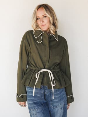 A relaxed but intentional outfit — an olive utility jacket with cream contrast piping and a drawstring waist pairs with lived-in blue jeans, shot against a soft neutral wall with an effortlessly cool, grounded energy.