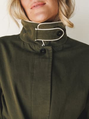 A close-up detail shot of an olive utility jacket with a structured funnel collar, cream drawstring tie, and dark button closures — the kind of jacket that makes a Tuesday feel intentional.