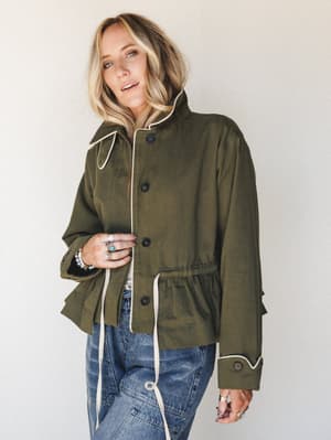 An olive utility jacket with cream contrast piping and a ruffled peplum hem is styled over blue jeans — the kind of piece that makes a casual outfit feel like it had a whole plan.